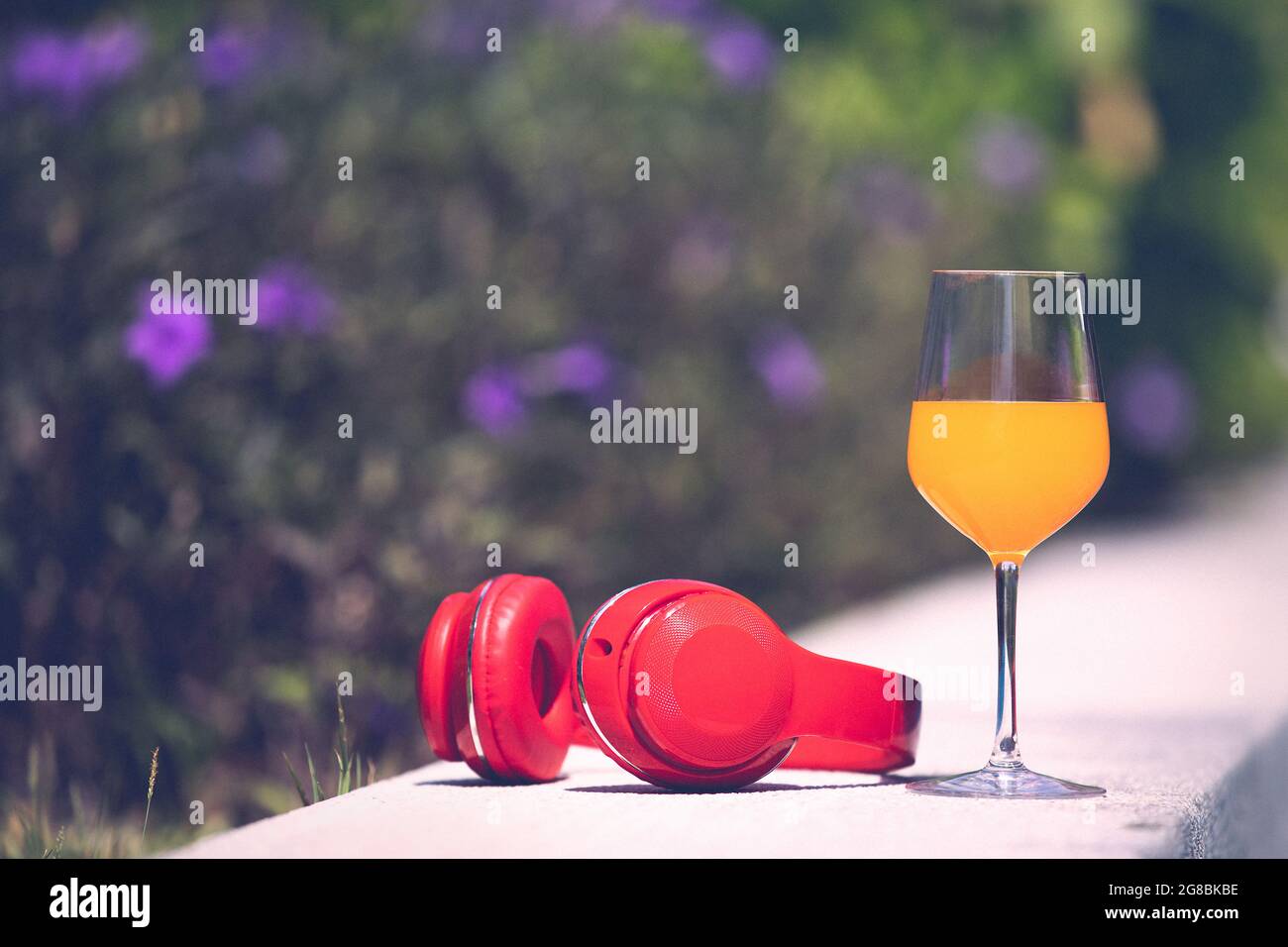 Glass of orange juice and red headphone on edge of swimming pool with blur flower and green tree in background. Copyspace on the left, process in vint Stock Photo