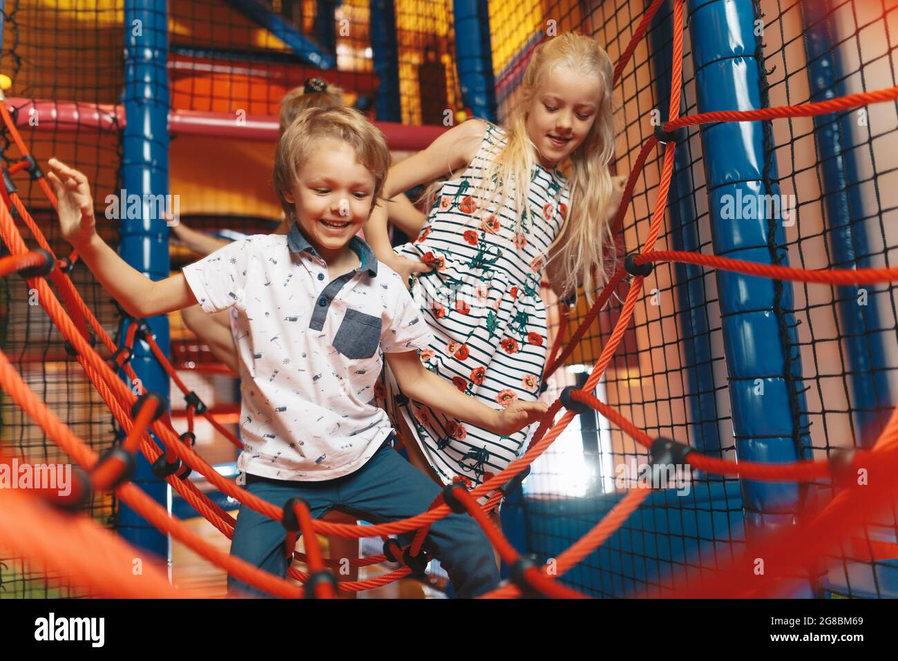 Happy group of siblings playing together on indoor playground. Excited kids playing together on net ropes. Cute school kids playing on colorful playgr Stock Photo