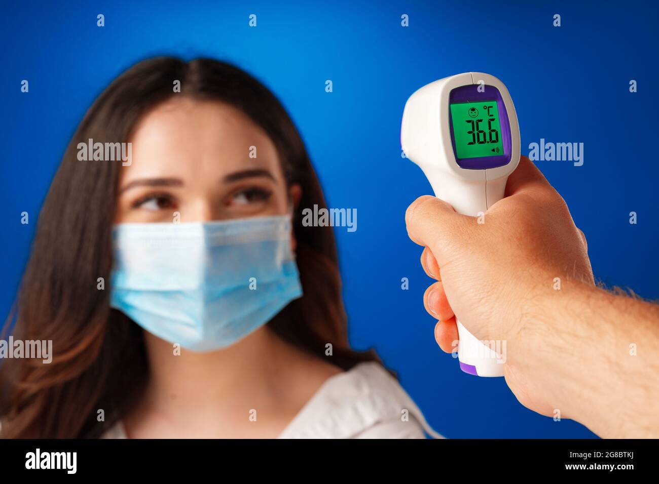 brunette woman getting temperature screening with infrared thermometer against blue background 2G8BTKJ
