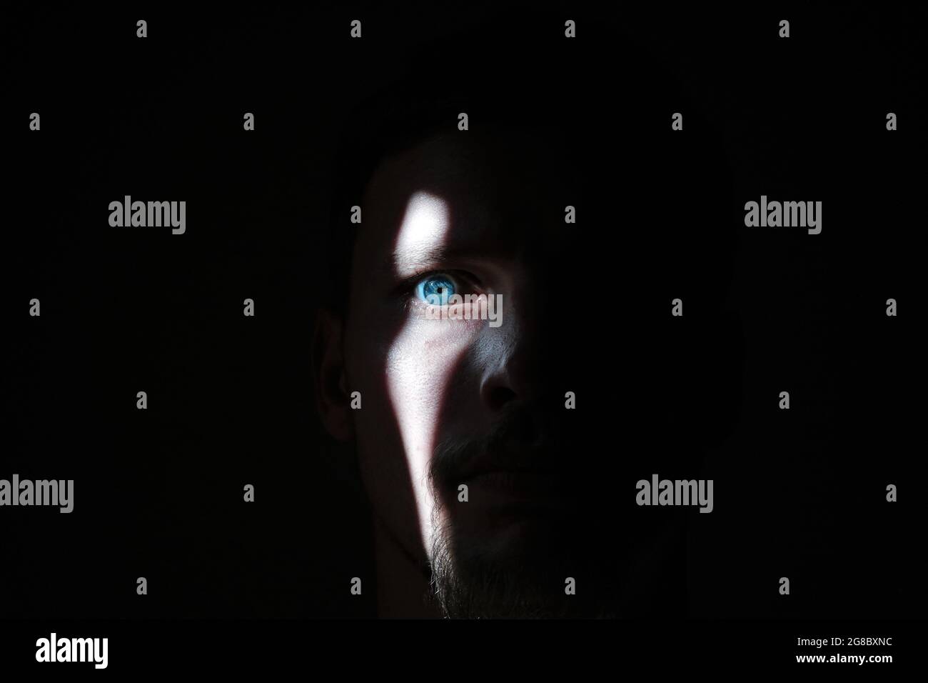 A sliver of bright light reveals a mans face and blue eye, everything else is shrouded in darkness. Stock Photo