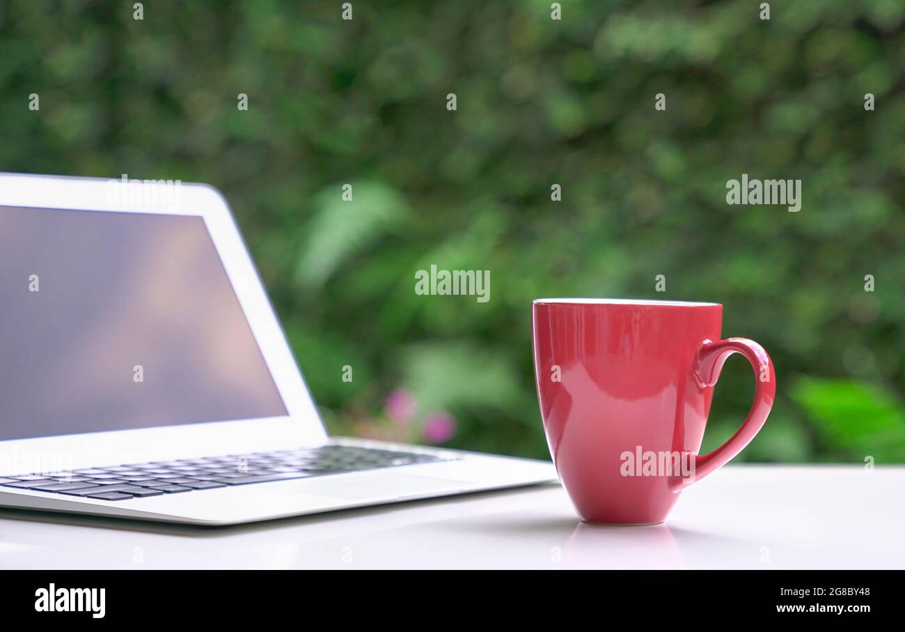 Good morning with coffee in red cup with computer laptop, green garden bokeh background. Copy space. Stock Photo