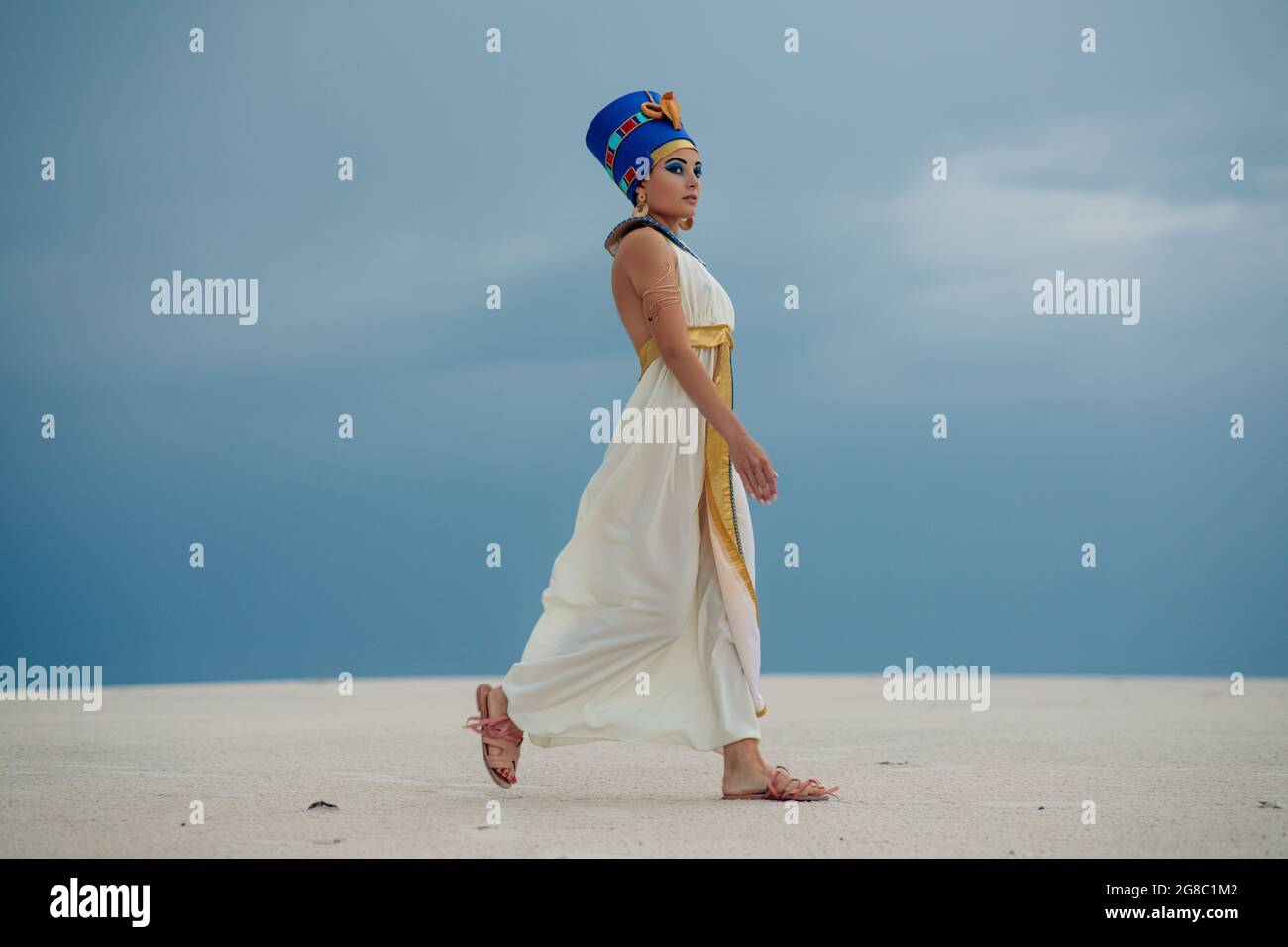 Young woman in image of ancient egyptian queen Nefertiti walks through sandy desert against background of blue sky. Stock Photo