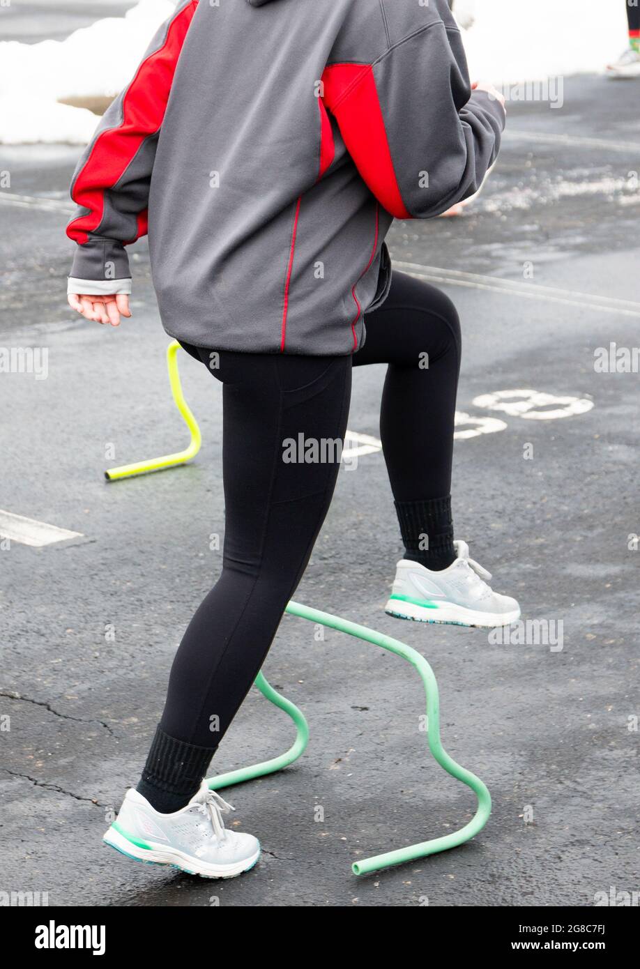 A high school girl is stepping and running over small yellow and green hurdles in a parking lot during track and field practice. Stock Photo