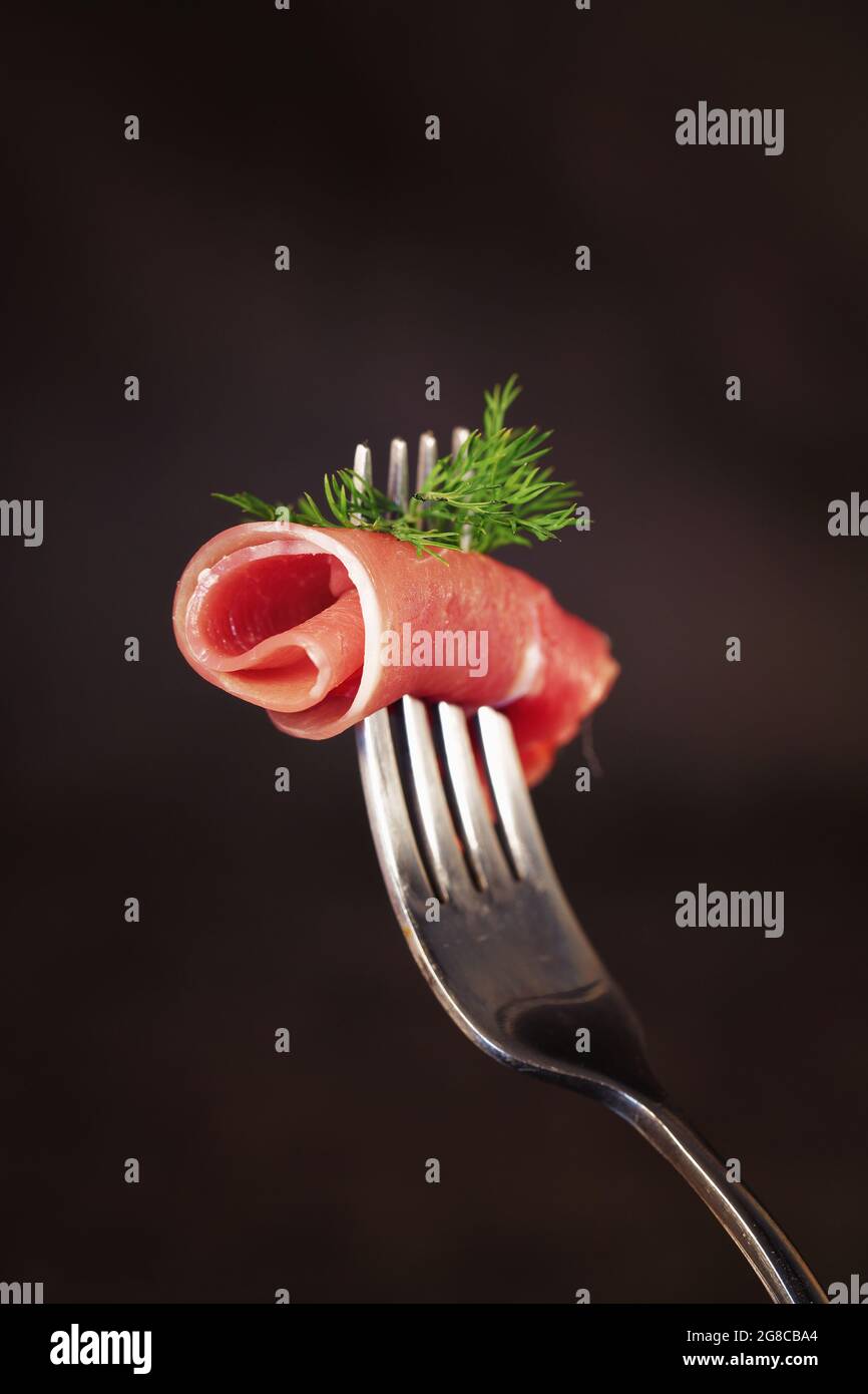 slice of dry ham rolled on a fork, isolated on black background Stock Photo