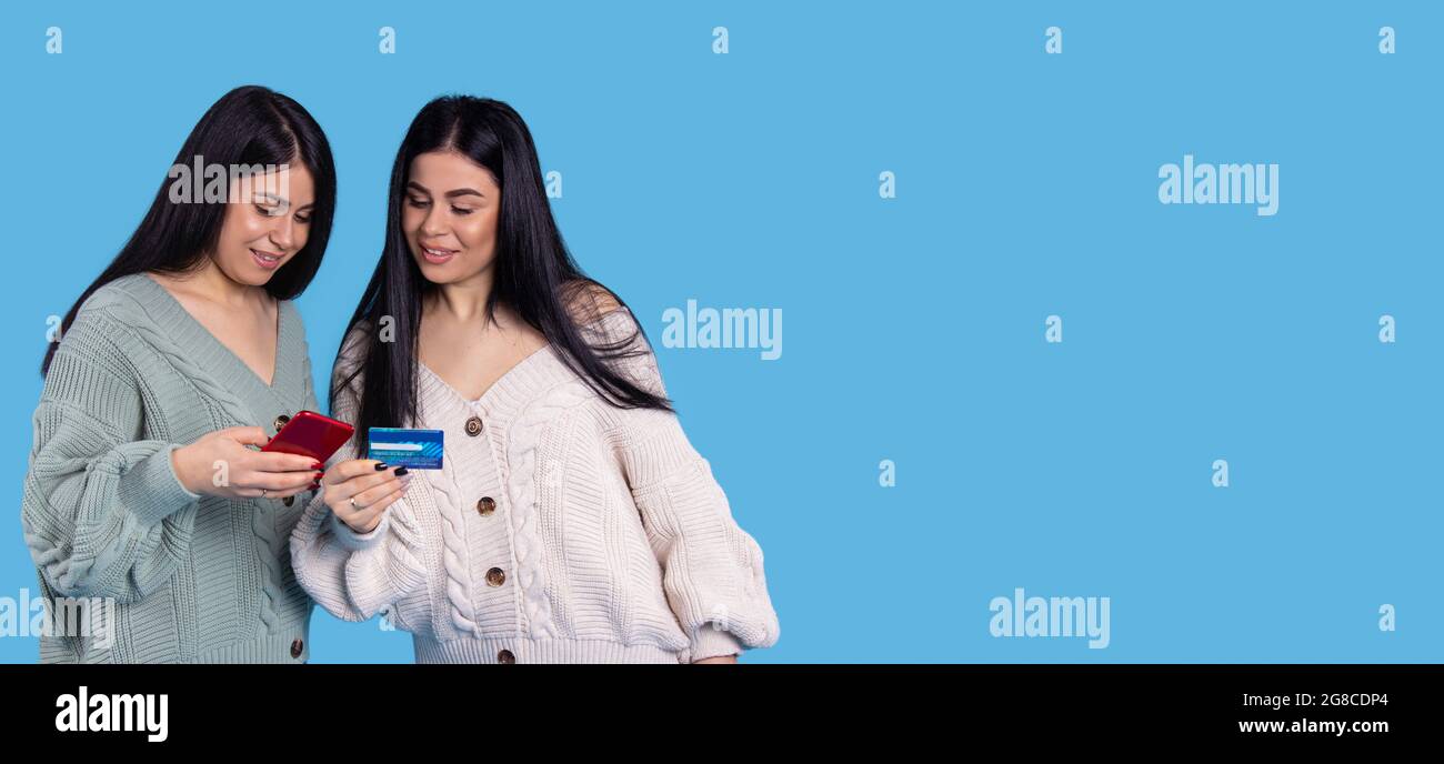 Banner, long format. Twins girls in knitted sweaters are holding cellphone and enter data from the card. Stock Photo