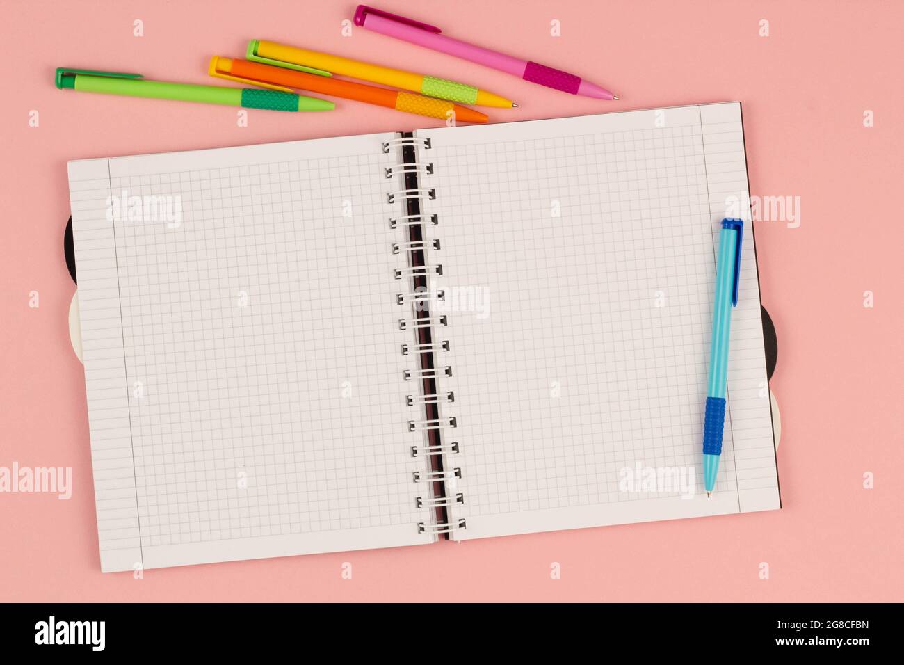 Open school squared notebook and colorful pens on the pink background. Blank white sheet of paper book on the table. Office supplies on the desktop Stock Photo