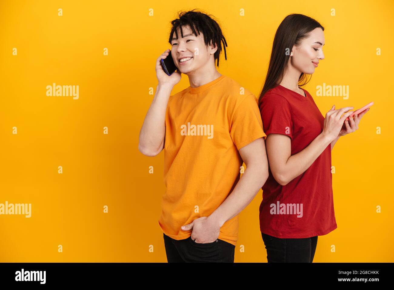 Young multiracial couple smiling while using and talking on cellphones isolated over yellow wall Stock Photo
