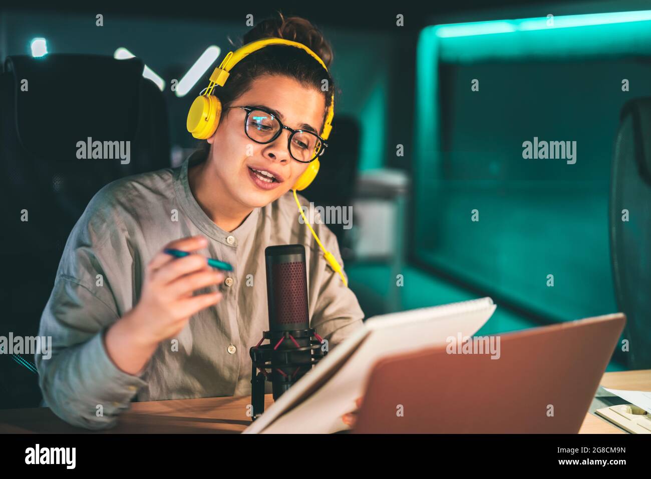 Young woman recording an audio podcast in a modern studio, wearing headphones and hipster glasses, speaking to the microphone and holding a notepad Stock Photo