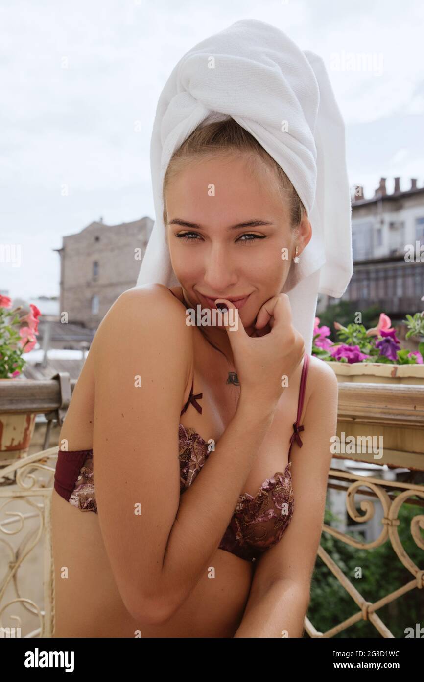 Girl in lingerie with white towel draped around her hair sits on a balcony in Little Paris (Odessa) on a sunny day,looking at us with thumb near mouth Stock Photo