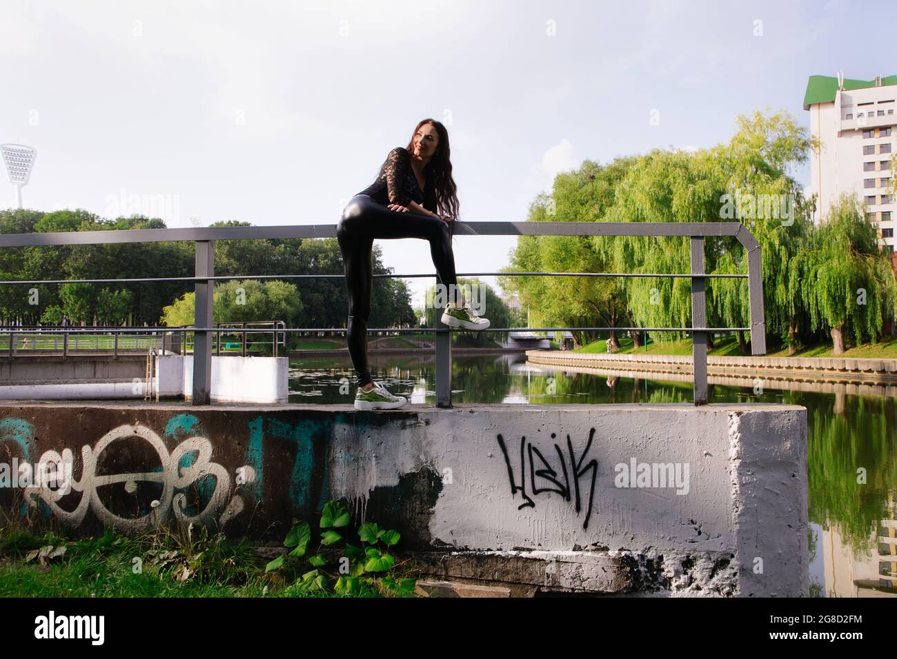Pretty female with long dark hair stands on stone object in water; she wears black clothes (transparent top & shiny tight trousers) & golden sneakers Stock Photo