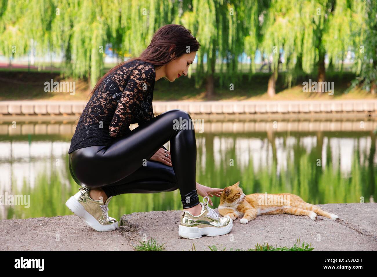 Pretty long-legged girl squats near water & pets an orange cat; she wears black clothes (transparent top & shiny tight leggings) and golden sneakers Stock Photo