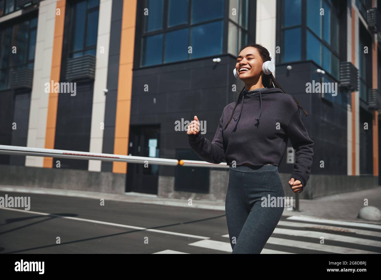 Smiling joyous sportswoman in wireless headphones and activewear jogging ahead Stock Photo