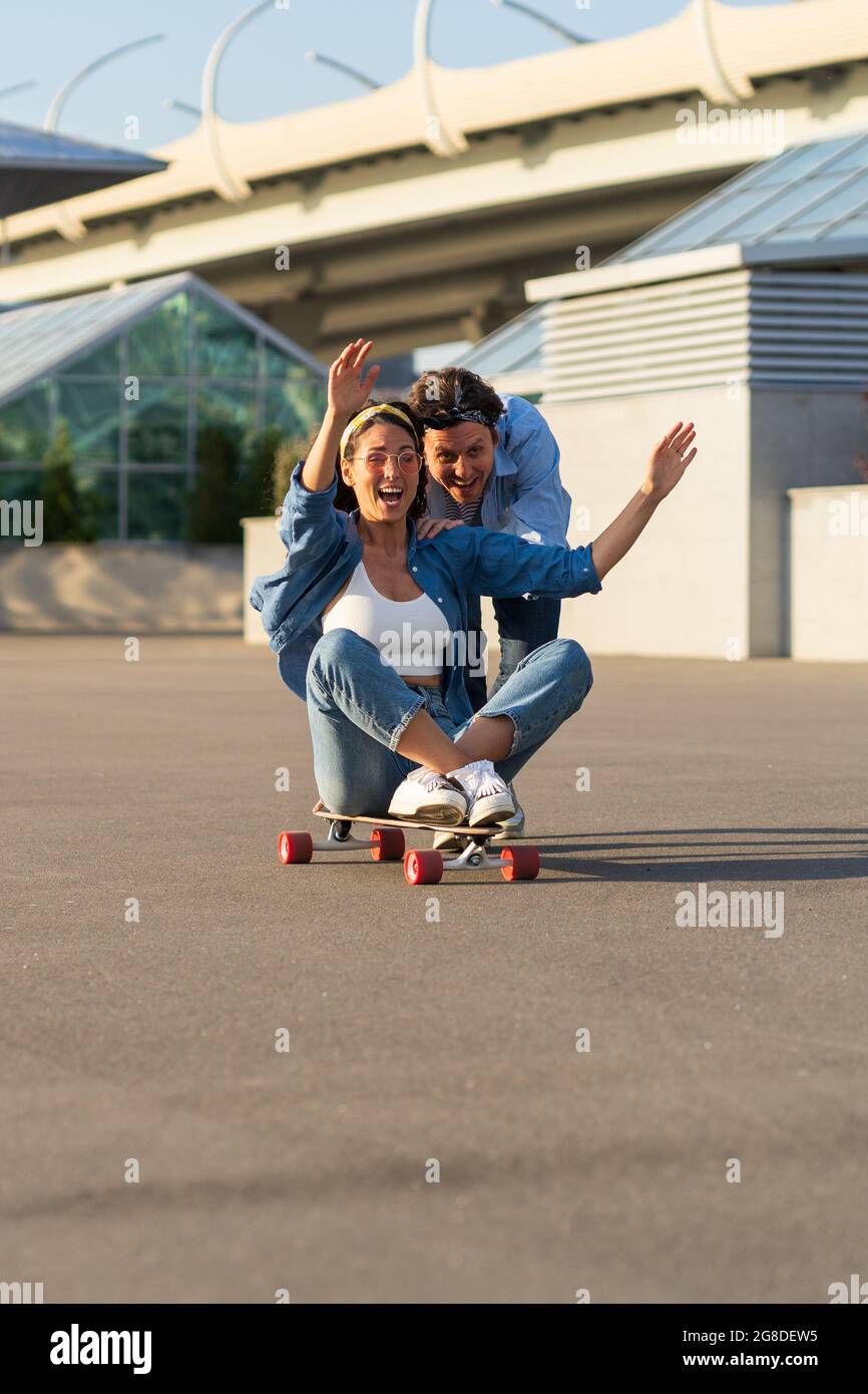Couple have fun on longboard: male push back of joyful female sitting on skateboard and laughing Stock Photo