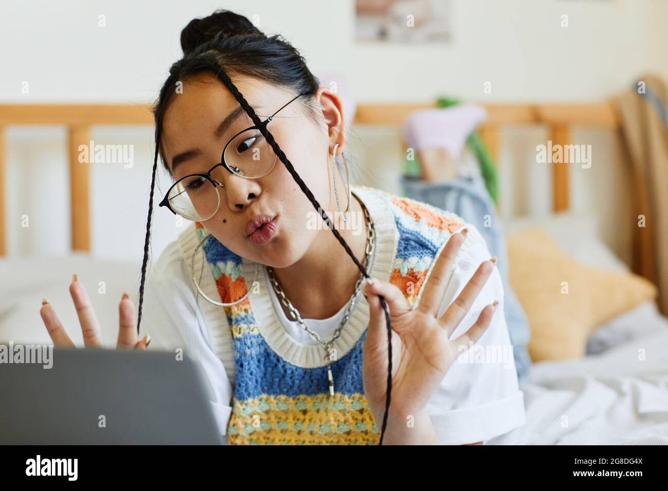 portrait of teenage asian girl using laptop on bed in cozy room interior and making cute faces to camera by video chat or livestream 2G8DG4X