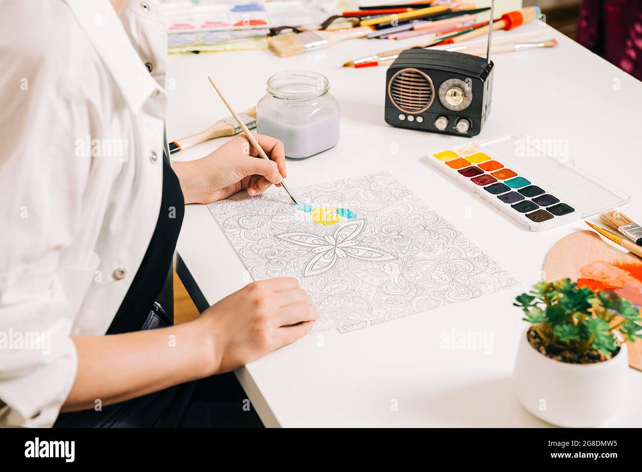 Young woman coloring page antistress at table indoors, mental wellbeing and art therapy. Woman paints a sketch, meditative process of coloring pages. Stock Photo