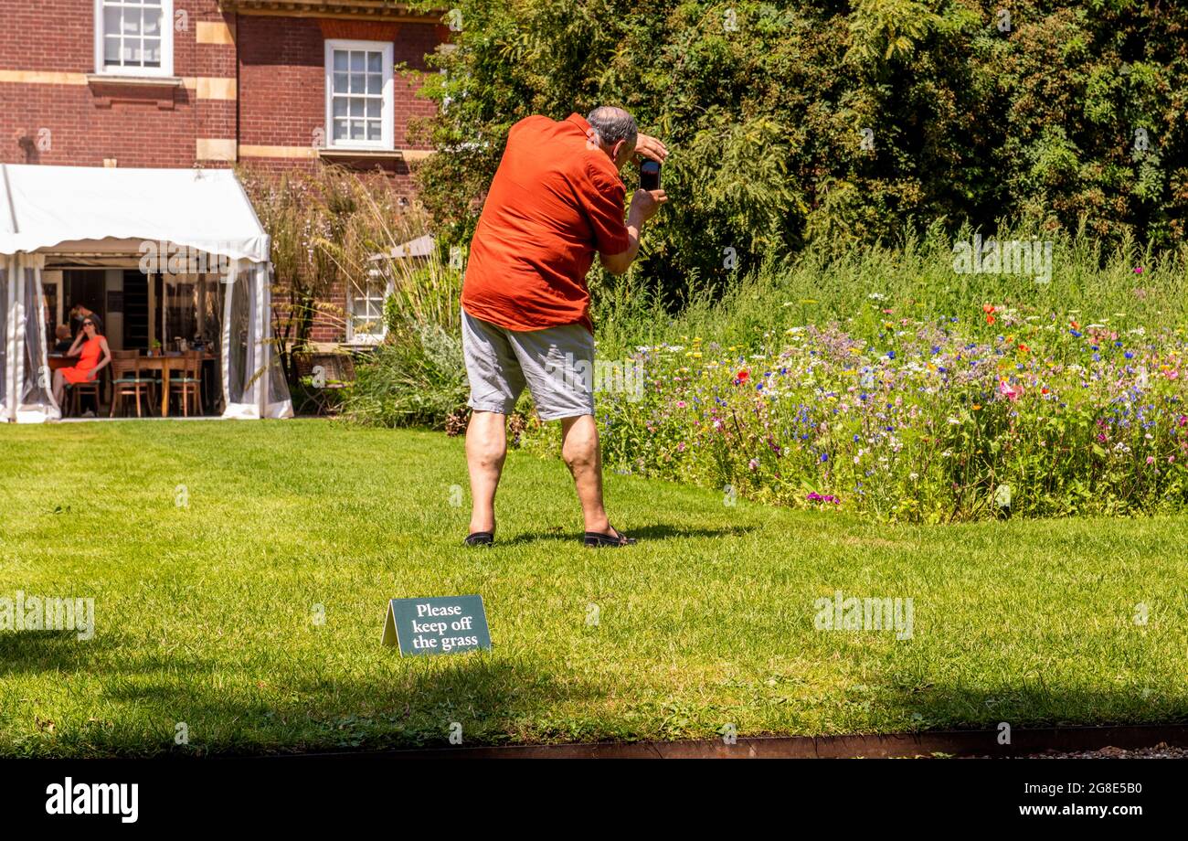 A man in an orange shirt is taking a picture while stepping on a lawn inches away from the sign 'Please keep off the grass'. Stock Photo