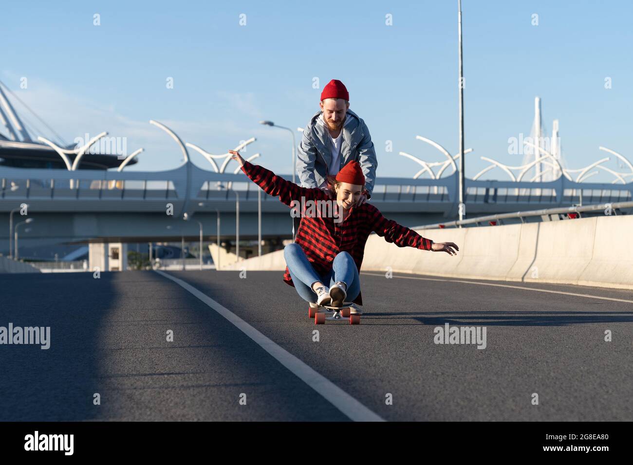 Casual couple on longboard: man teaching girl to balance on skateboard push her back down empty road Stock Photo