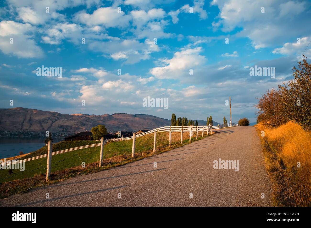 white fence near the road, farmhouse, lake, village and hills in the background. Blue sky with white clouds. Stock Photo