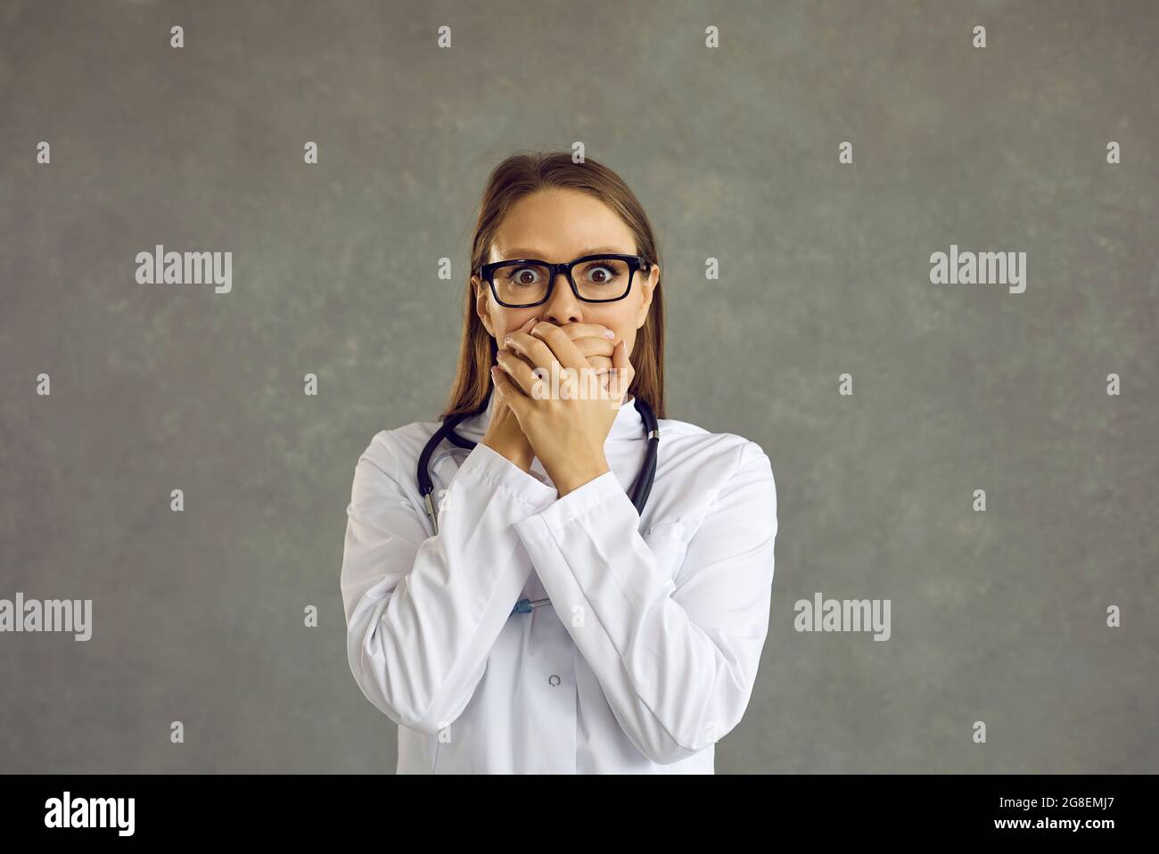 Worried female doctor with a frightened expression covering her mouth on a gray background. Stock Photo