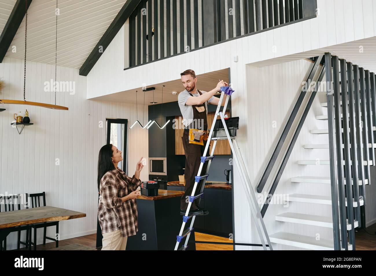 Young worker on stepladder talking to female client while fixing alarm system Stock Photo