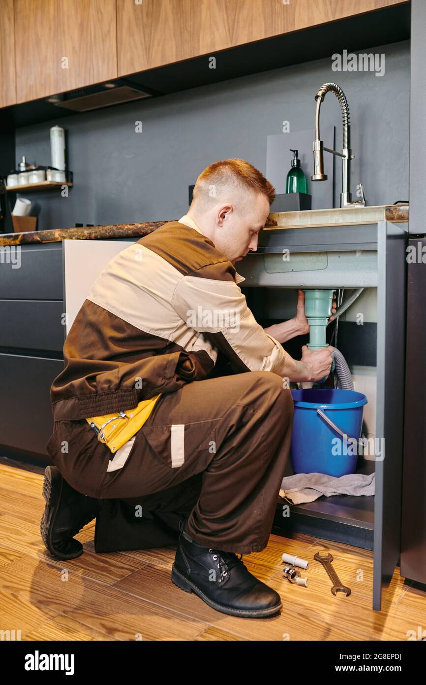 Modern plumber in workwear changing pipe under sink in the kitchen