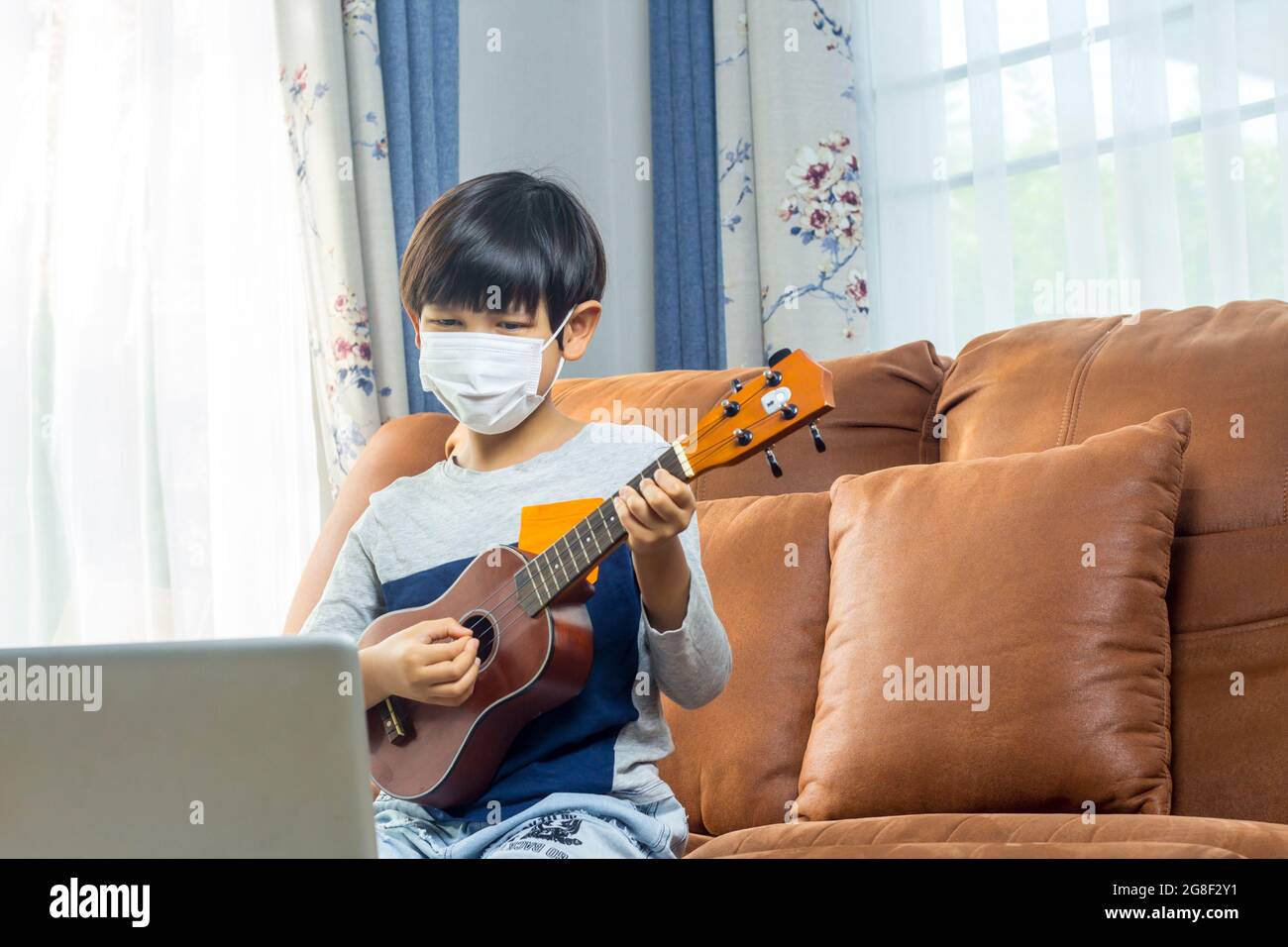 A boy wearing a face mask plays ukulele in front of a laptop Stock