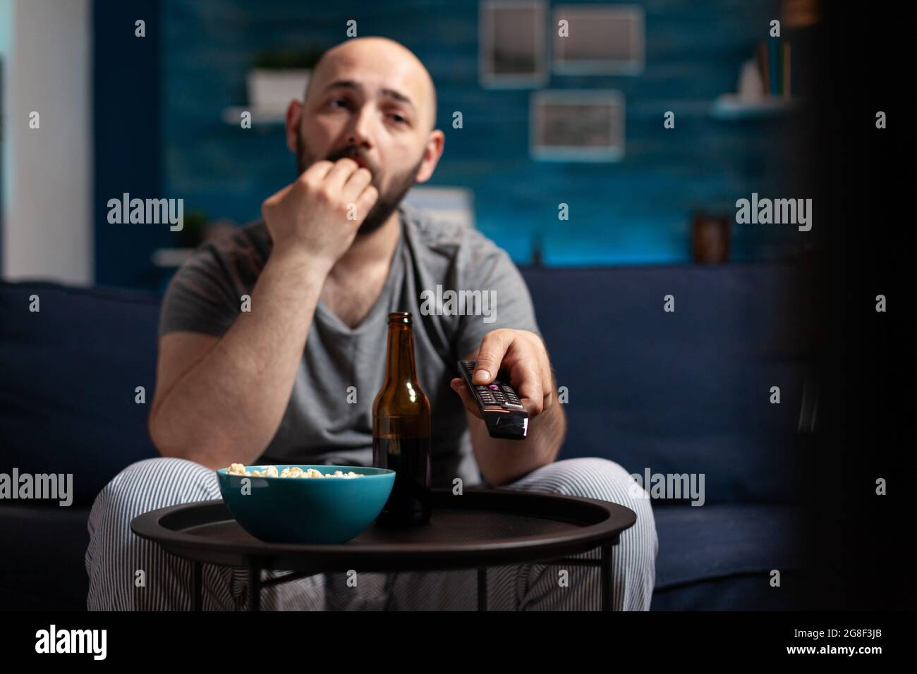 Scared shocked man looking at horror thriller movie at tv, eating popcorn trembled with fright sitting on comfortable sofa. Concentrated alone male watching tv late night enjoying free time Stock Photo