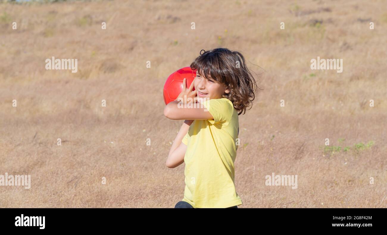 smiling young boy with long hair and yellow t-shirt plays throwing his red ball in the field in summer on a sunny day Stock Photo