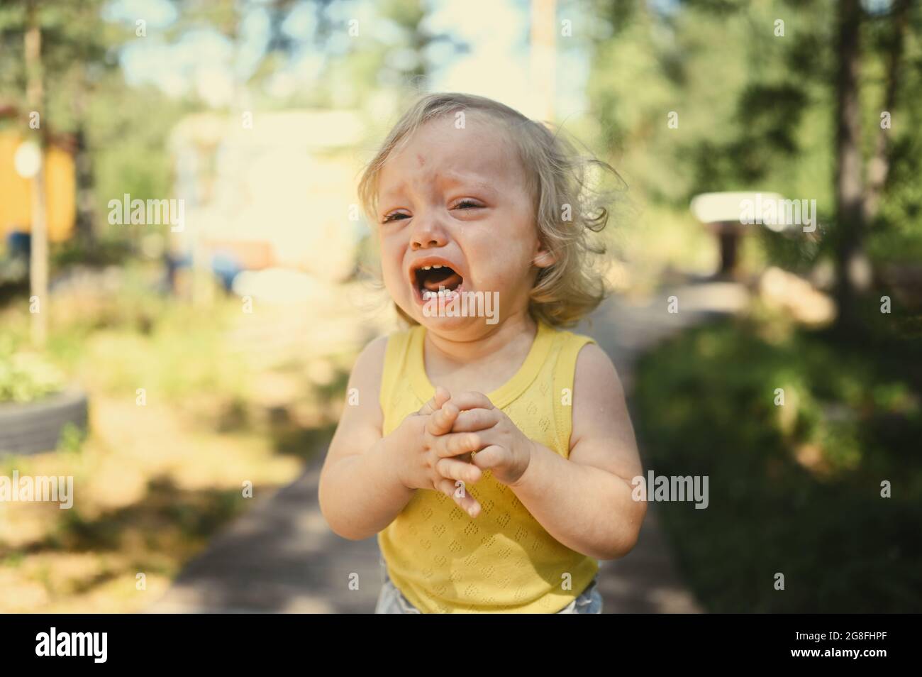 Close up portrait of little funny cute blonde girl child toddler in yellow bodysuit crying outside at summer. Childish tantrum. Healthy childhood concept Stock Photo