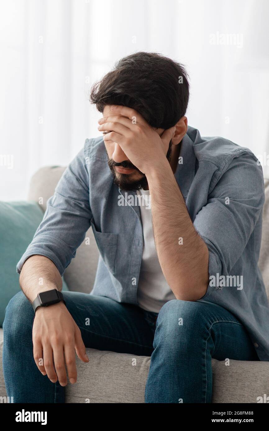 Male depression concept. Desperate arab man touching his forehead, having problems, sitting on sofa at home, crop Stock Photo