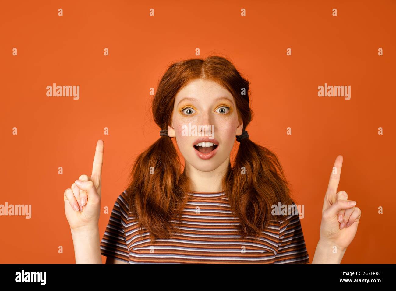 Ginger shocked woman with tails pointing fingers upward isolated over orange background Stock Photo