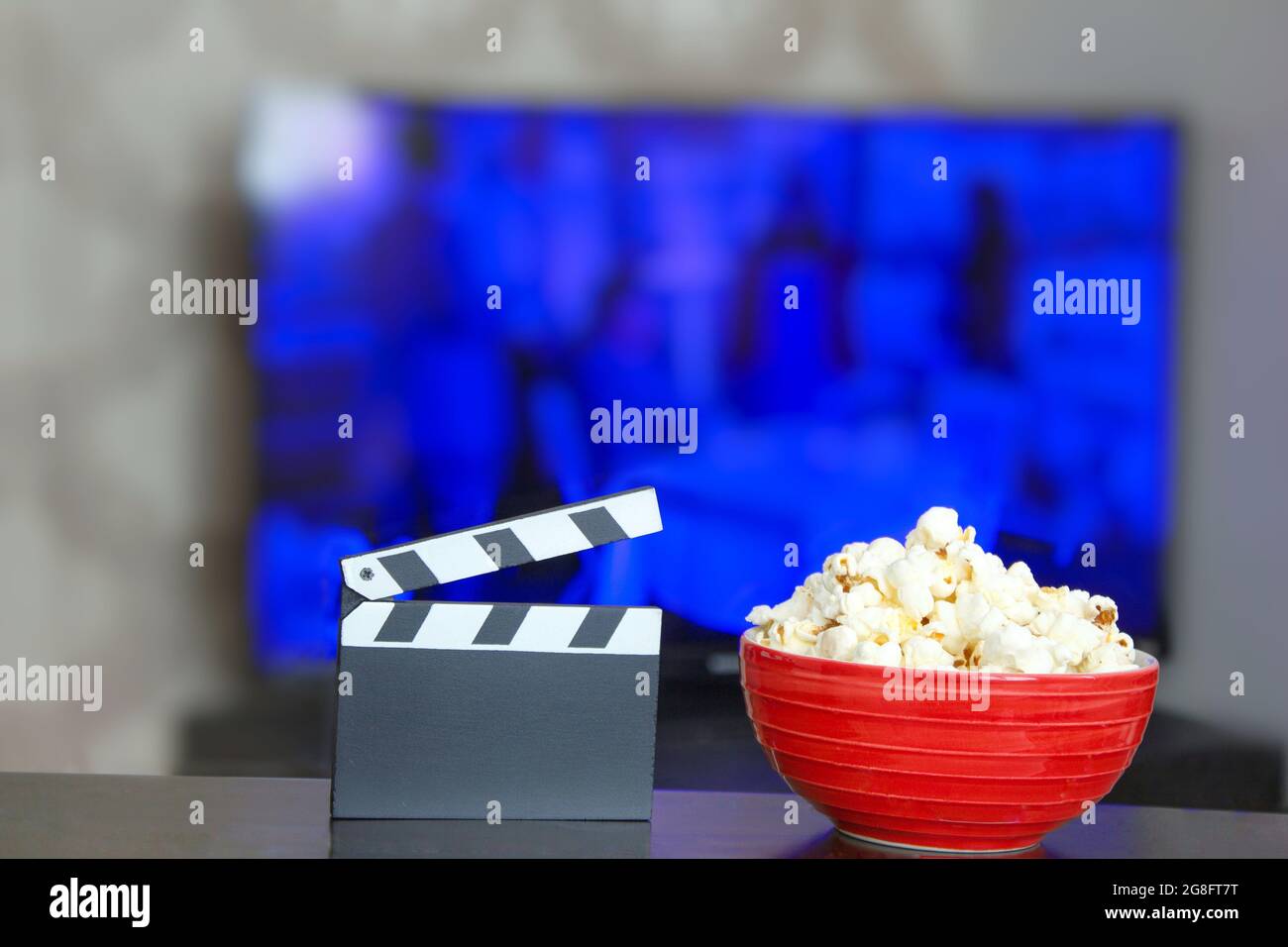 Black movie clapper board and a bowl of popcorn, blurry television in background. Entertainment industry background photo. Movie time. Watching tv. Stock Photo