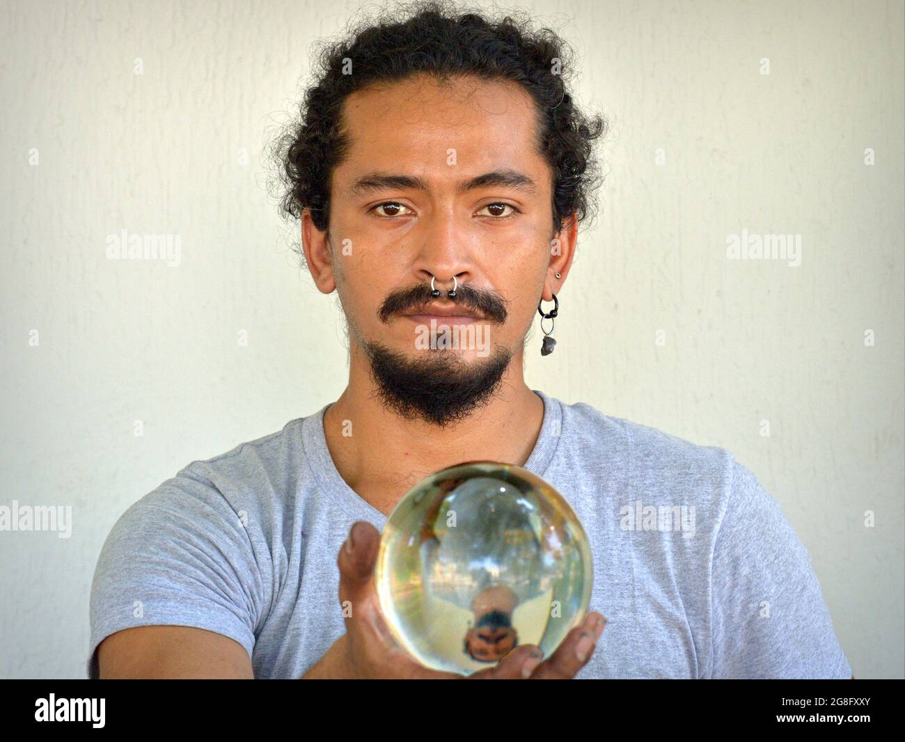Handsome young Hispanic Latino man with nose ring holds a transparent crystal ball with his mirror image in his right hand and looks at the viewer. Stock Photo