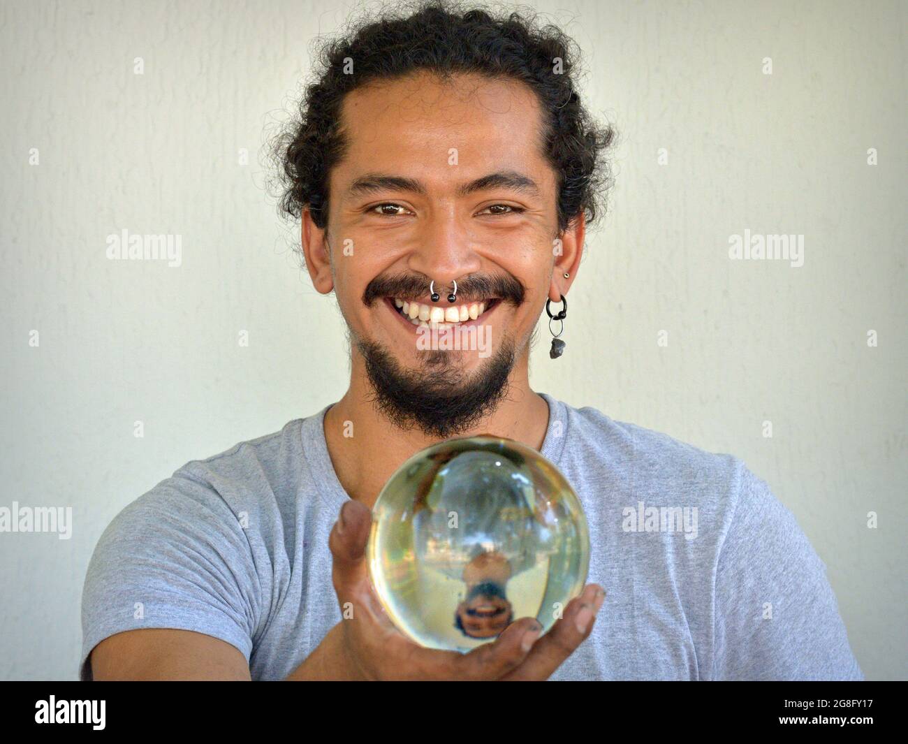 Handsome young Hispanic Latino man with nose ring holds a transparent crystal ball with his mirror image in his right hand and smiles at the viewer. Stock Photo