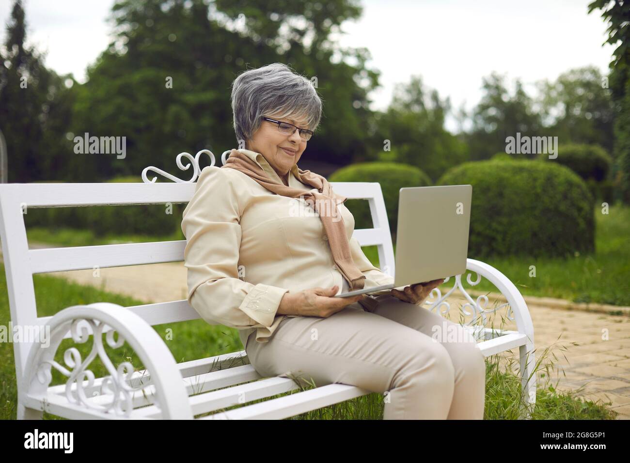 Happy senior lady using laptop sitting on bench relaxing in park outdoors Stock Photo