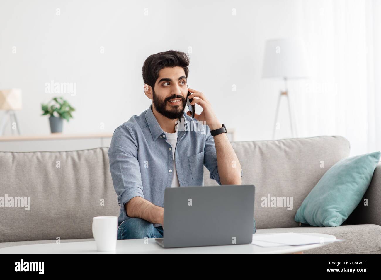 Freelance lifestyle. Arab man using laptop and talking on cellphone, working while sitting on sofa in living room Stock Photo
