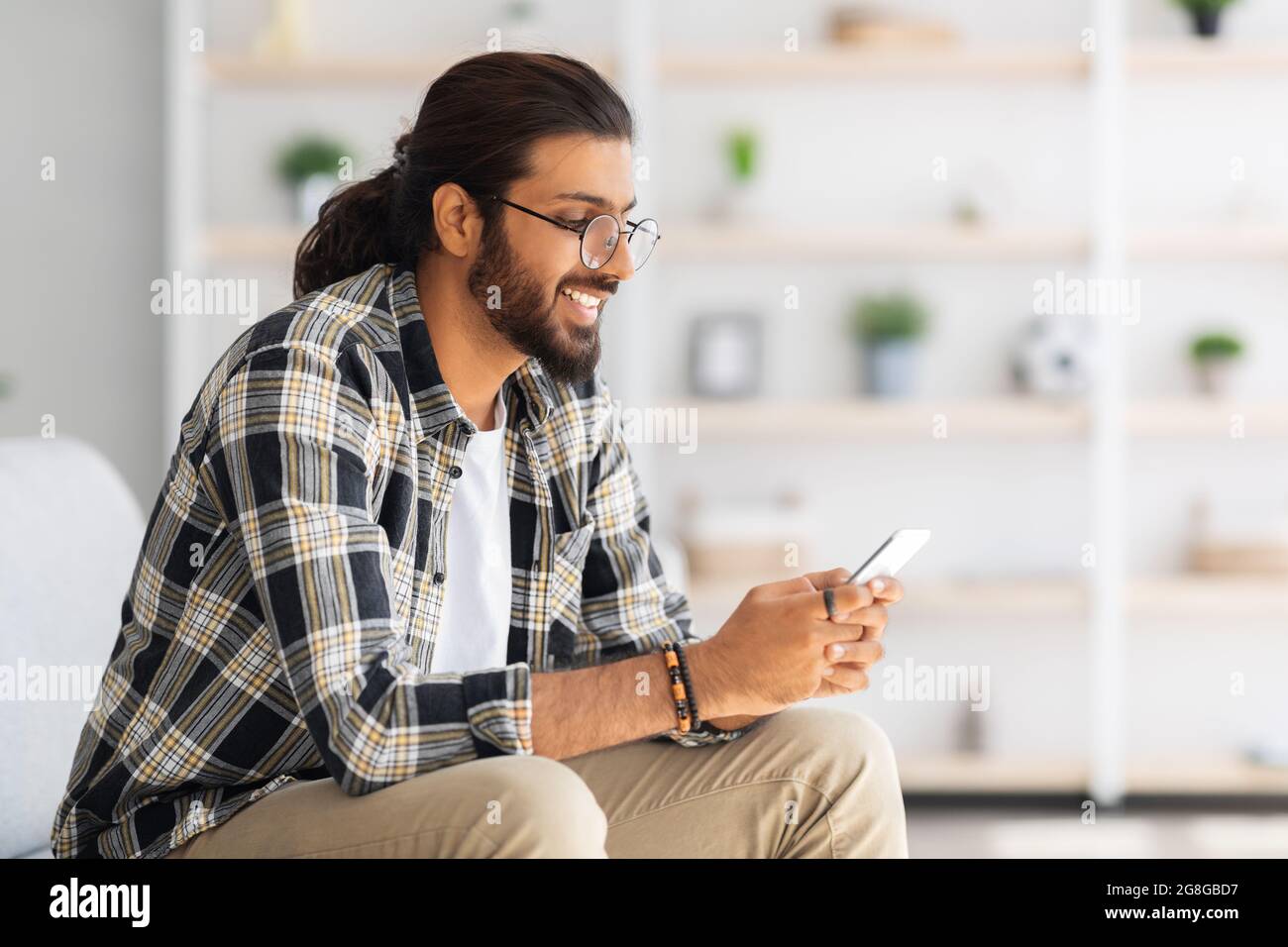 Positive long-haired middle-eastern guy sitting on couch, using cellphone Stock Photo