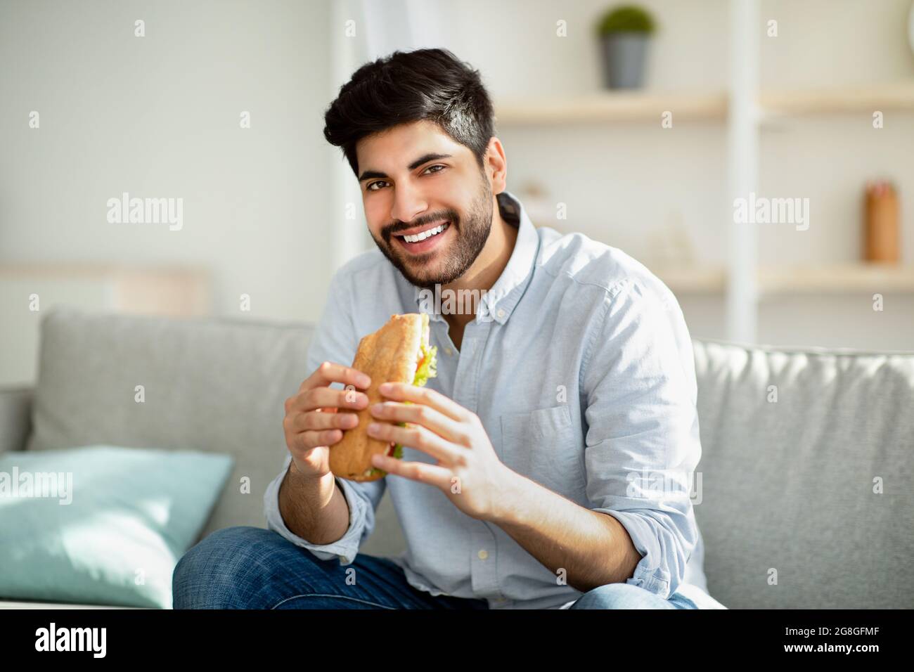 Cheat meal day. Happy arab man eating tasty sandwich and smiling to camera, sitting on sofa in living room at home Stock Photo