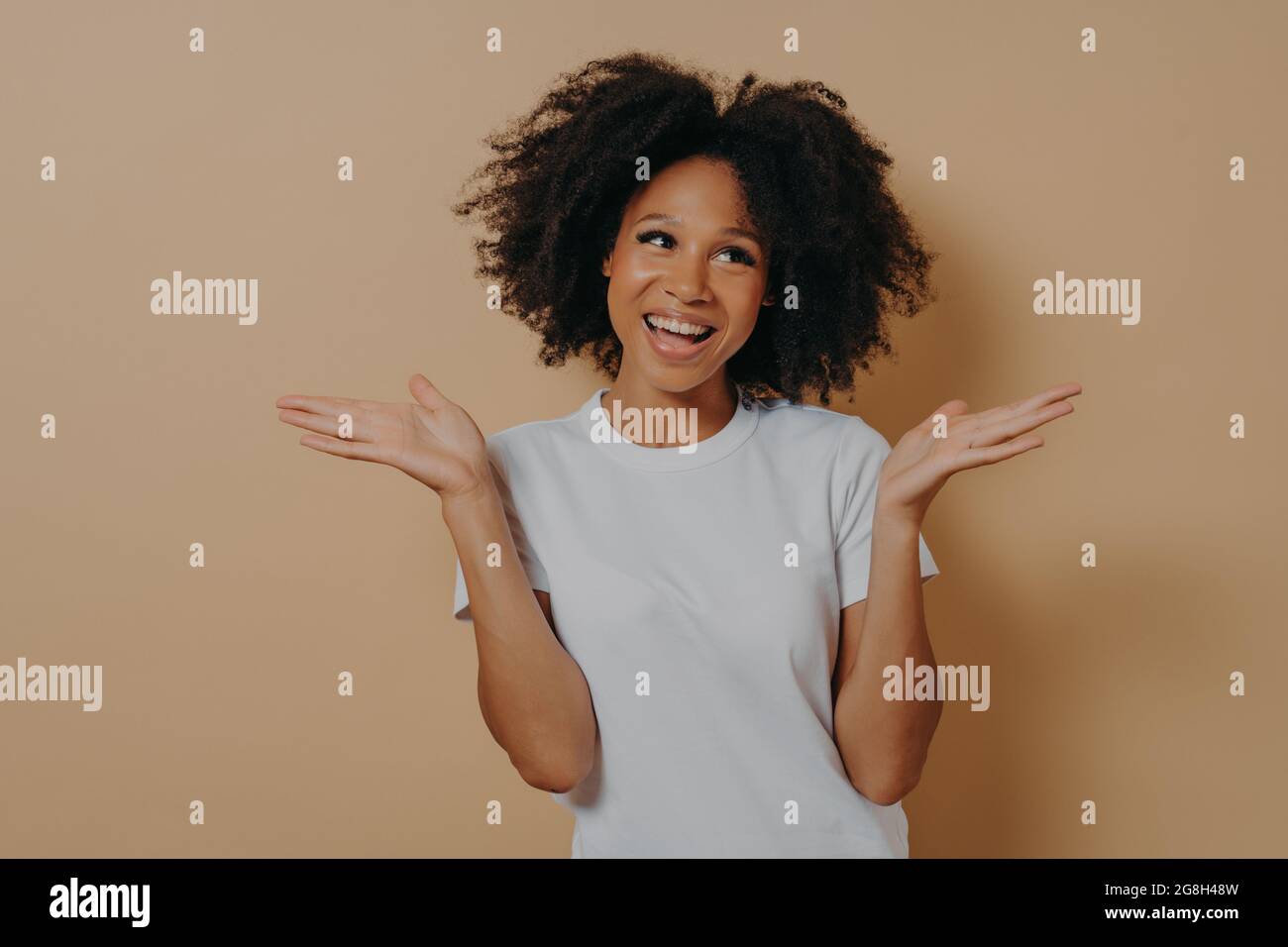 Young smiling mixed race woman raising hands with hesitation, isolated on beige background Stock Photo