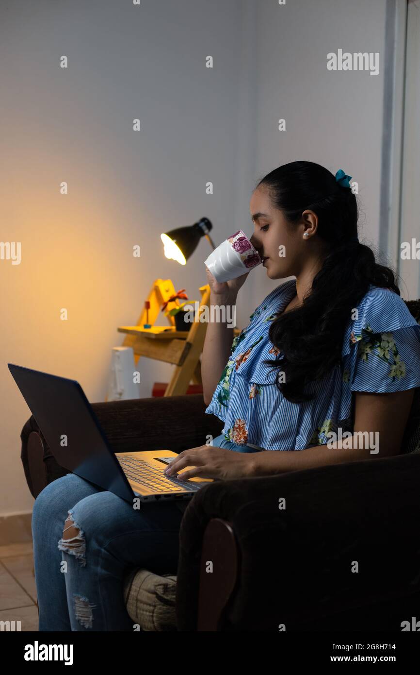 Young Hispanic female drinking coffee while working from home sitting on the armchair Stock Photo