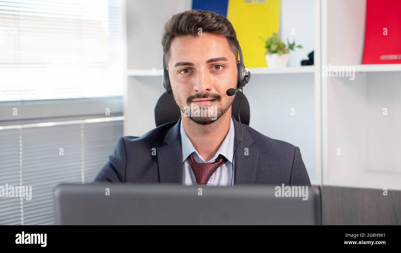 Portrait of businessman, call center agent wearing microphone headset working with laptop in office.  Man in suit consulting client on phone in custom Stock Photo