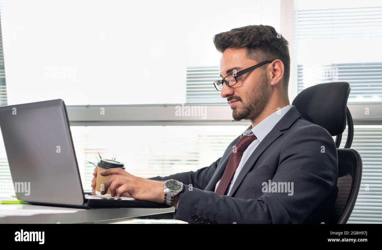 Portrait of happy businessman in formal clothing working with laptop in office. Cheerful, relaxed man in suit working while drinking coffee or tea. Stock Photo