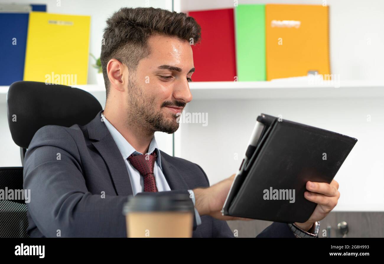Cheerful young professional businessman in suit using digital tablet while sitting at desk in modern office. Stock Photo