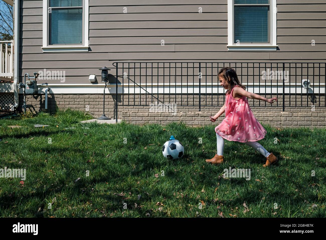 Little girl in pink dress kicking soccer ball in back yard Stock Photo