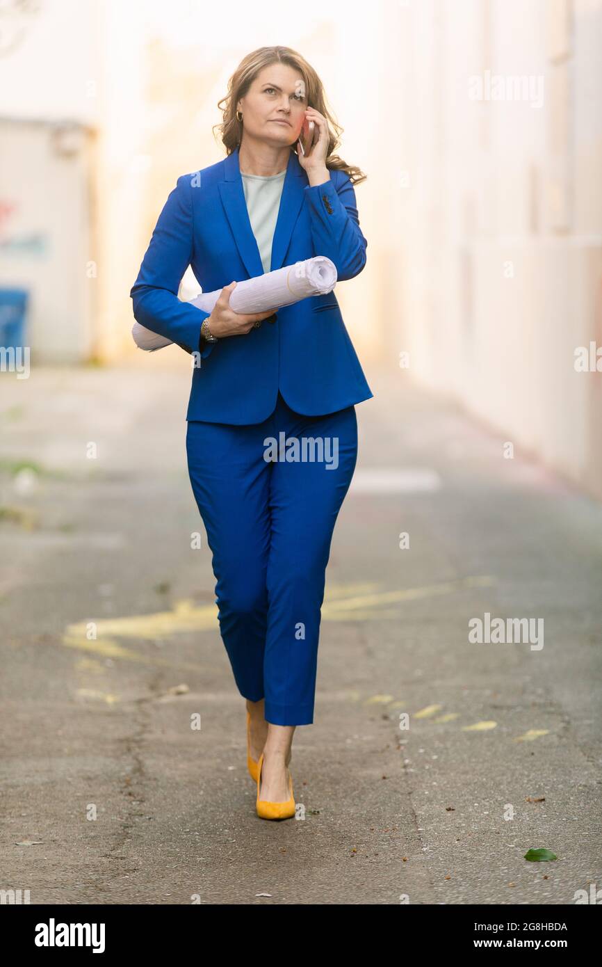 Full body of middle aged female project manager in elegant blue suit with rolled paper in hands answering phone call while walking on urban street Stock Photo
