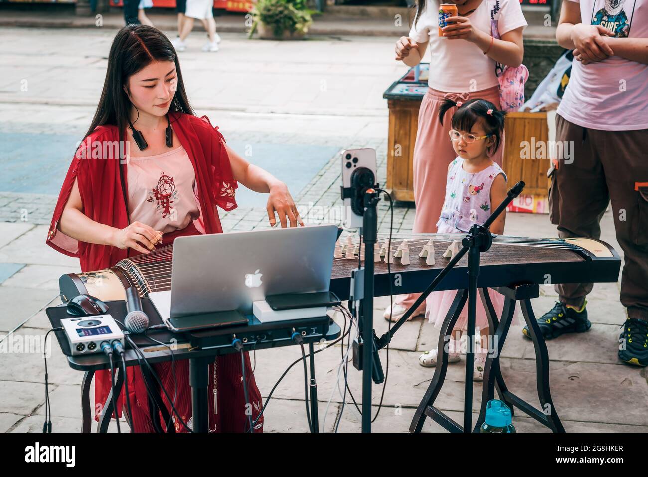 Luodai, Chengdu, Sichuan province, China - June 27, 2021 : Little girl watching a young chinese woman playing guzheng in the street Stock Photo