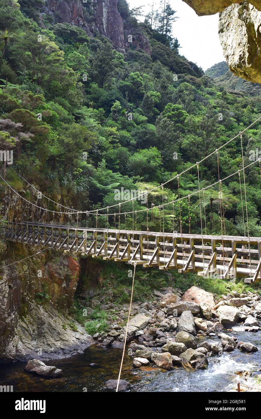 Suspension bridge connecting two cliffs over shallow river with many boulders at opening into valley with native bush. Stock Photo