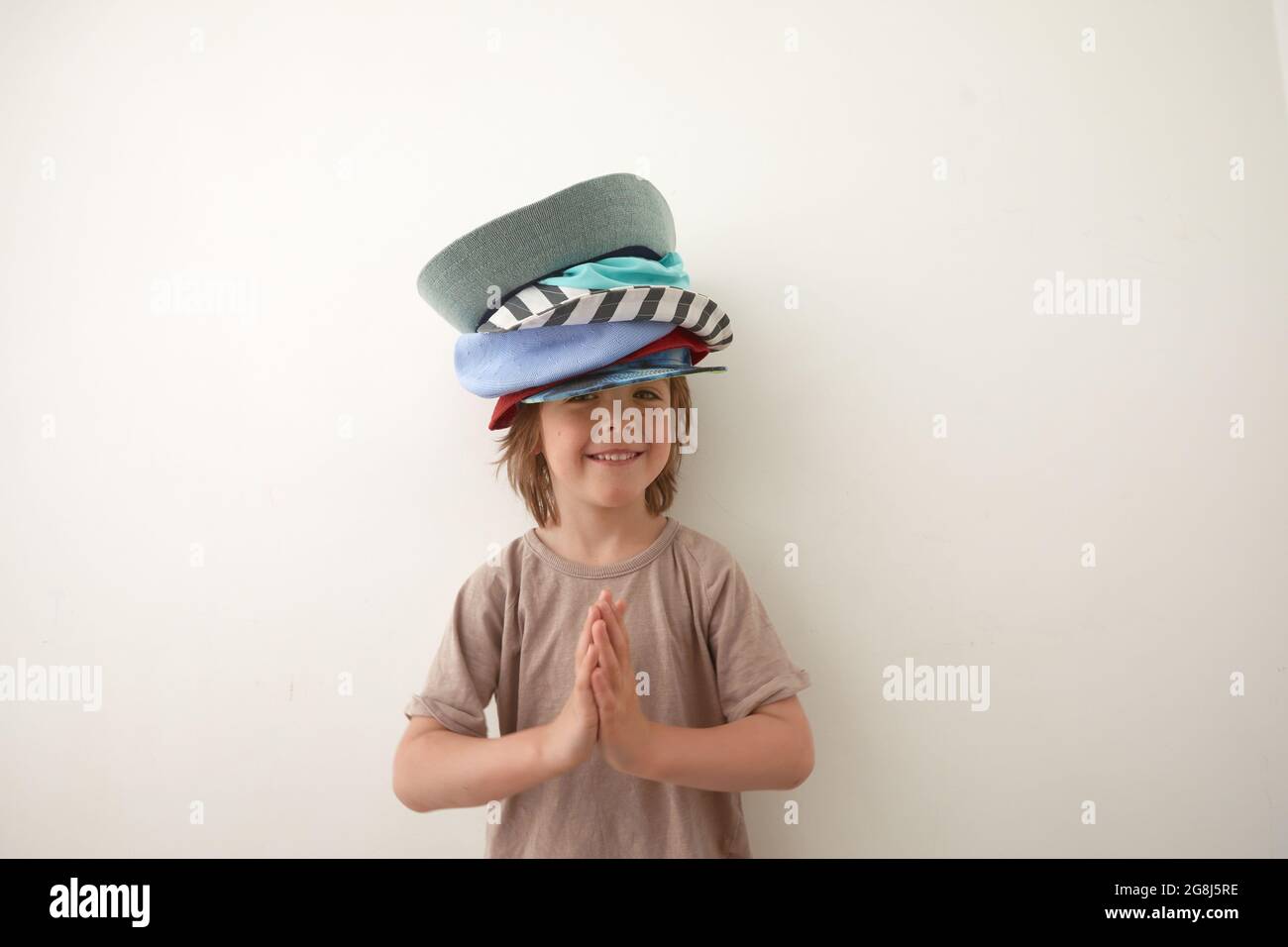 Smiling boy wearing t shirt and various headwear standing against white background and looking at camera. Child's palms are folded at the chest in gra Stock Photo