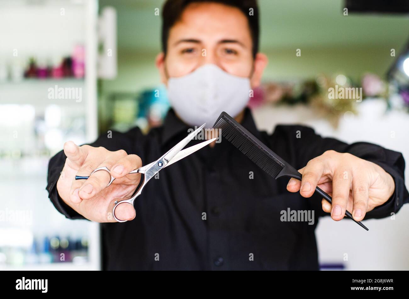 Portrait of an attractive barber, guy with safety mask, holding scissors and a comb in his hands, social distancing, preventive measures against the Stock Photo