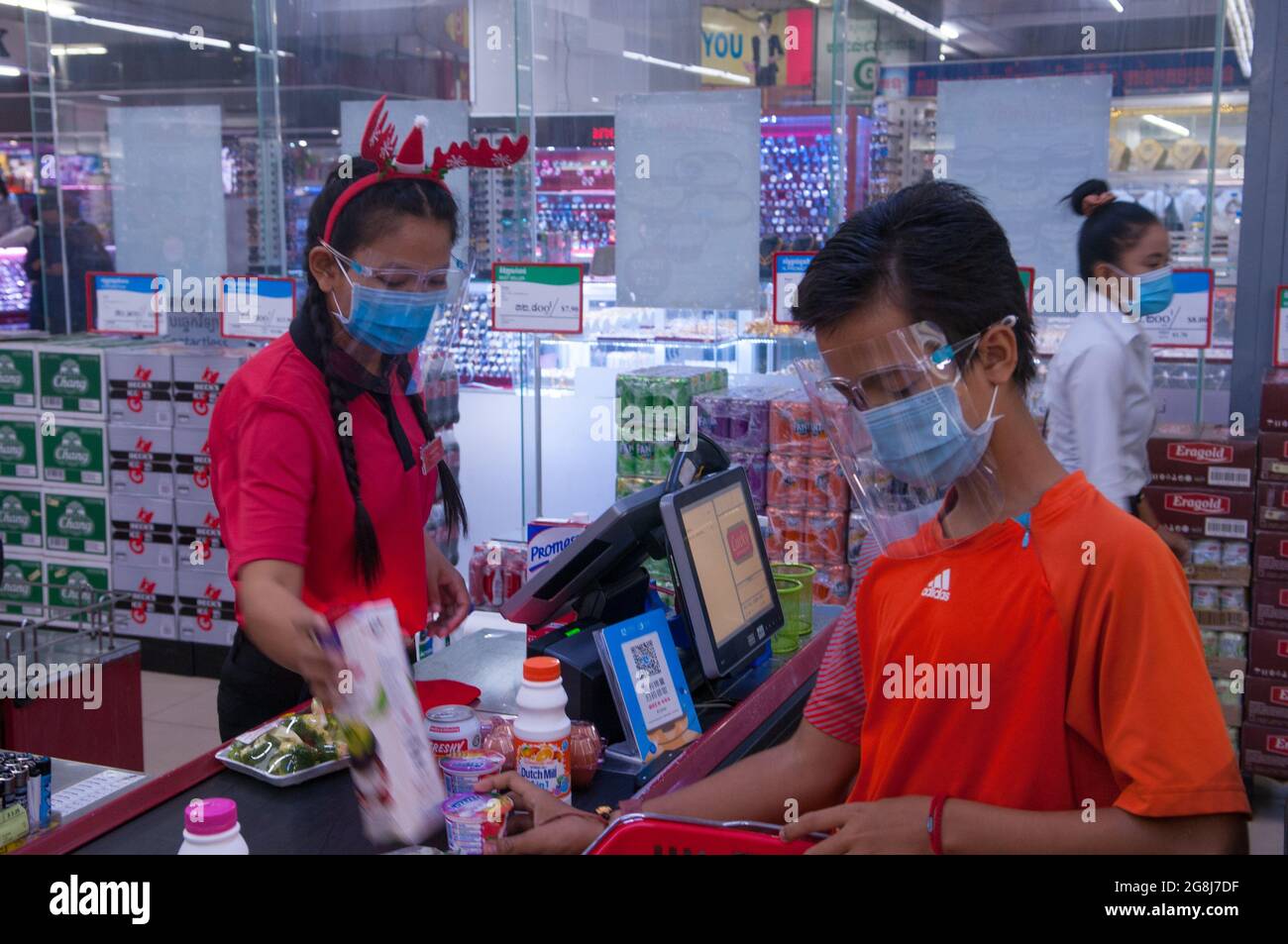 during the Christmas holidays with an outbreak of COVID - 19, a Cambodian female cashier, wearing a protective plastic face shield & mask, scans groceries at a grocery store checkout counter during the coronavirus pandemic. a mixed race teenage boy (Cambodian - American) loads his groceries onto the counter. Steung Meanchey, Phnom Penh, Cambodia. Dec. 23rd, 2020. © Kraig Lieb Stock Photo
